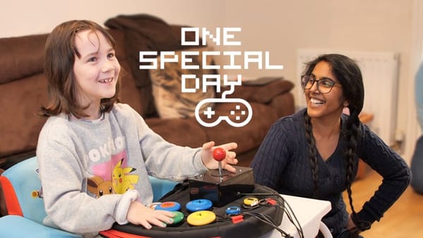 A child with a specially adapted video game controller smiling while playing something offscreen while an adult watches them, also smiling. 